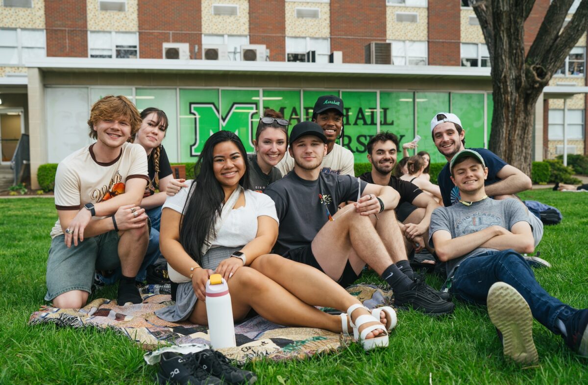 Marshall University students sit on campus on the grass, smiling together on a blanket. 