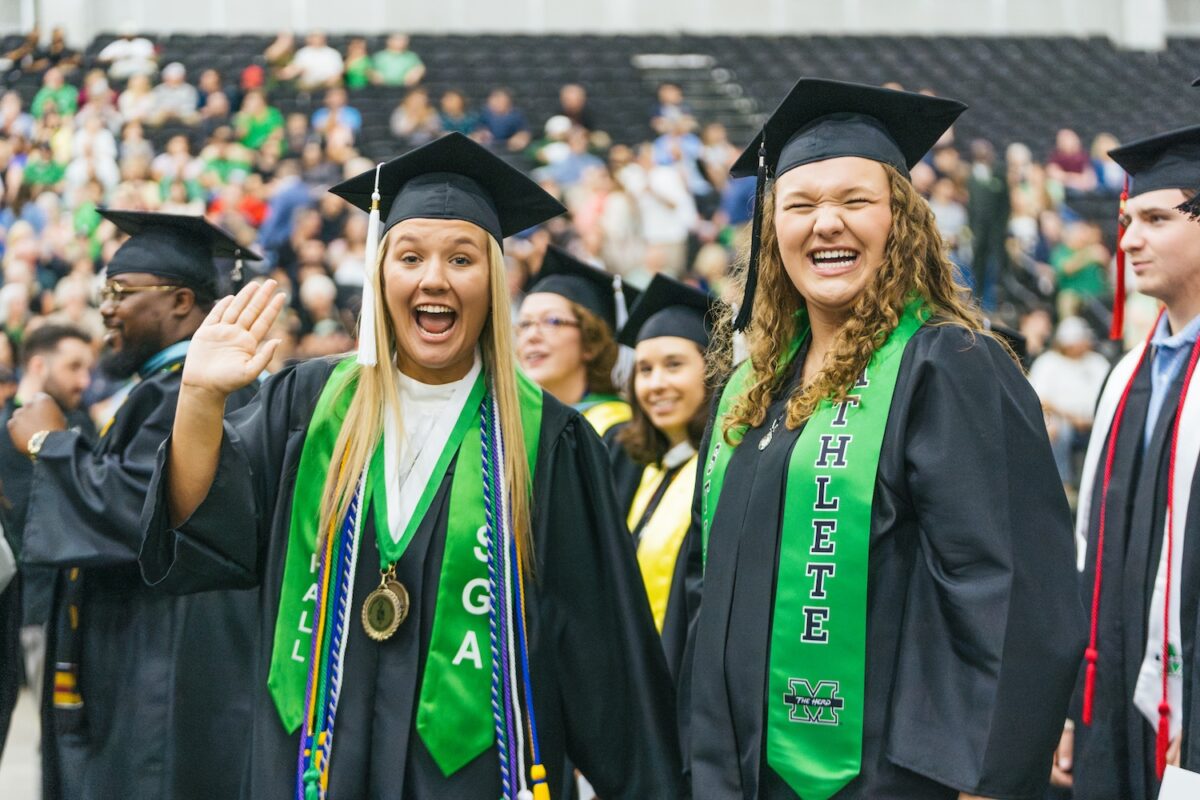 Two Marshall University students smile and wave at the camera in their caps and gowns.