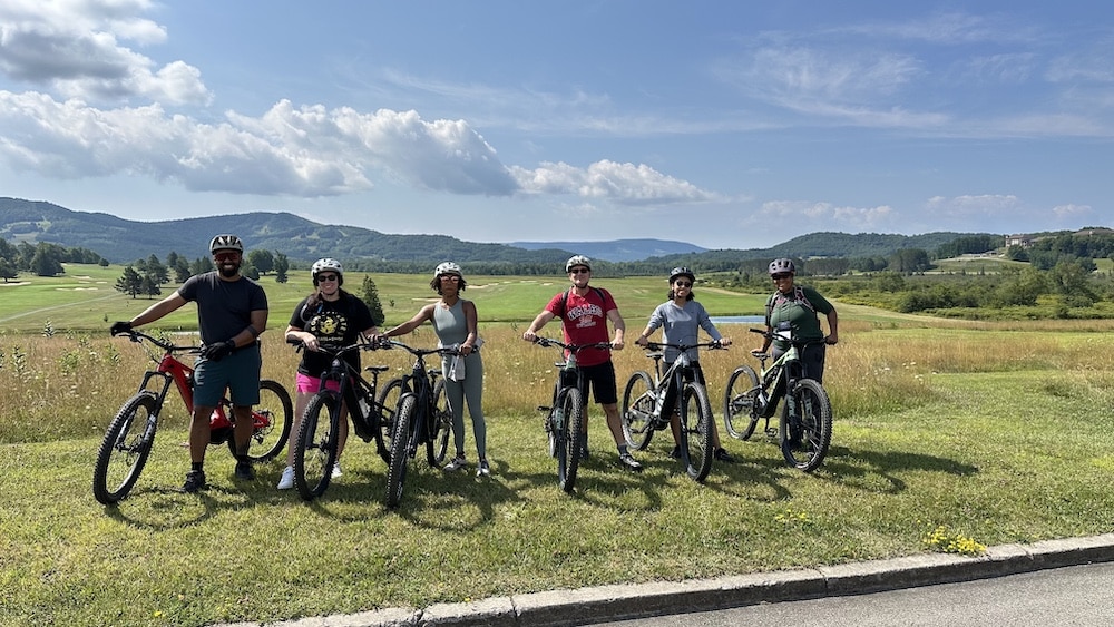 First Ascent members gather together on the side of a mountain biking trail, smiling with their bikes.
